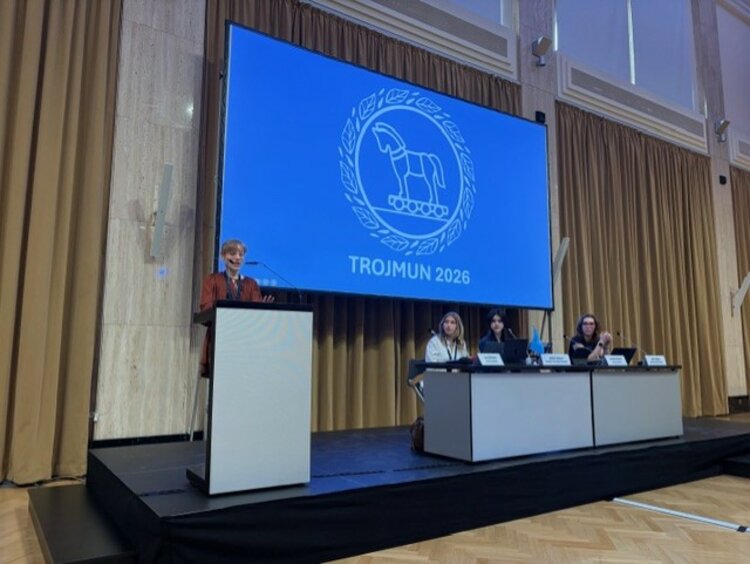 Modellsitzung der Vereinten Nationen TROJMUN 2026 Eine Person hält eine Rede an einem Rednerpult auf einer Bühne. Im Hintergrund zeigt eine große Leinwand das Logo und den Schriftzug „TROJMUN 2026“. Drei Personen sitzen an einem Tisch rechts auf der Bühne.