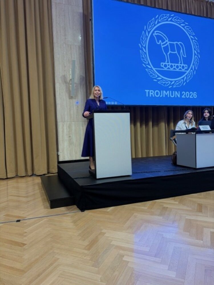 Modellsitzung der Vereinten Nationen TROJMUN 2026 Eine Frau in einem dunkelblauen Kleid steht an einem Rednerpult auf einer Bühne. Im Hintergrund ist eine große Leinwand mit dem Logo und dem Schriftzug „TROJMUN 2026“ zu sehen. Zwei weitere Personen sitzen an einem Tisch rechts auf der Bühne.