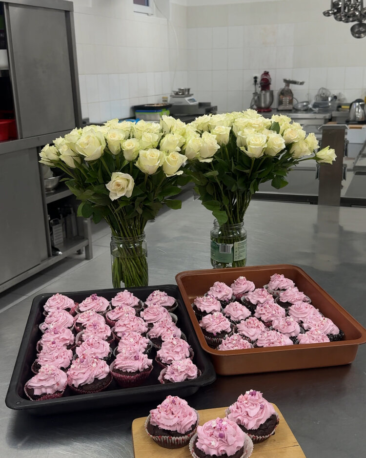 Valentinstag voller Freude und Hilfsbereitschaft Auf einem Tisch stehen zwei große Trays mit rosa verzierten Cupcakes. Dahinter sind zwei Vasen mit großen Sträußen weißer Rosen zu sehen. Im Hintergrund befindet sich eine Küche mit Arbeitsflächen und Geräten.