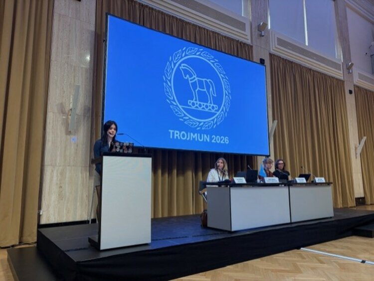 Modellsitzung der Vereinten Nationen TROJMUN 2026 Eine Person hält eine Rede an einem Rednerpult auf einer Bühne. Im Hintergrund zeigt eine große Leinwand das Logo und den Schriftzug „TROJMUN 2026“. Drei Personen sitzen an einem Tisch rechts auf der Bühne.