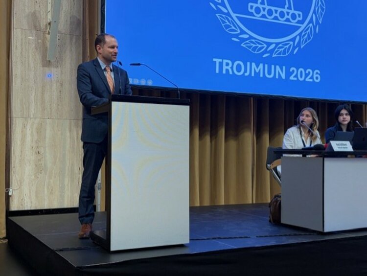 Modellsitzung der Vereinten Nationen TROJMUN 2026 Ein Mann in einem dunkelblauen Anzug und lachsfarbener Krawatte spricht an einem Rednerpult auf einer Bühne. Im Hintergrund ist eine Leinwand mit dem Logo und dem Schriftzug „TROJMUN 2026“ zu sehen. Zwei Personen sitzen an einem Tisch rechts auf der Bühne.