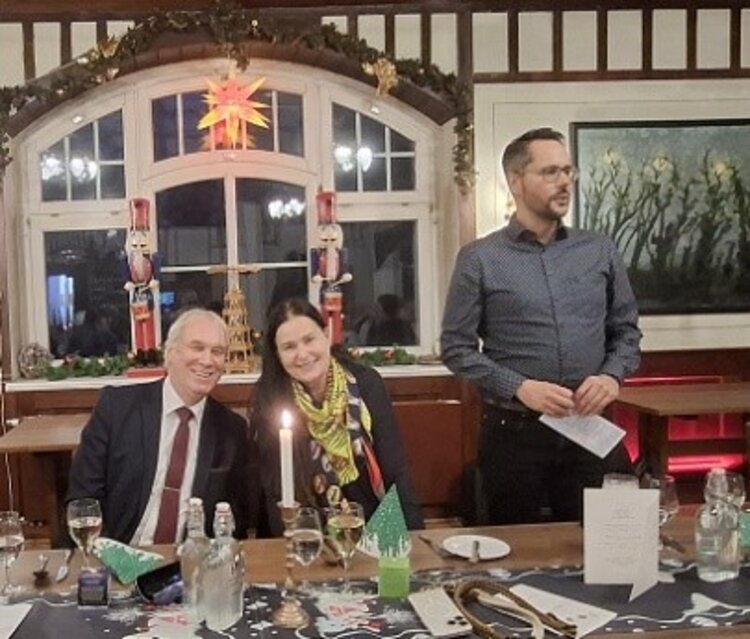 Räucherkerzen und ein Hauch von Weihnacht Drei Personen sitzen an einem festlich geschmückten Tisch im historischen Saal, zwei lächeln in die Kamera, während der Mann rechts stehend eine Ansprache hält; im Hintergrund ein großes Fenster mit Weihnachtsdeko.