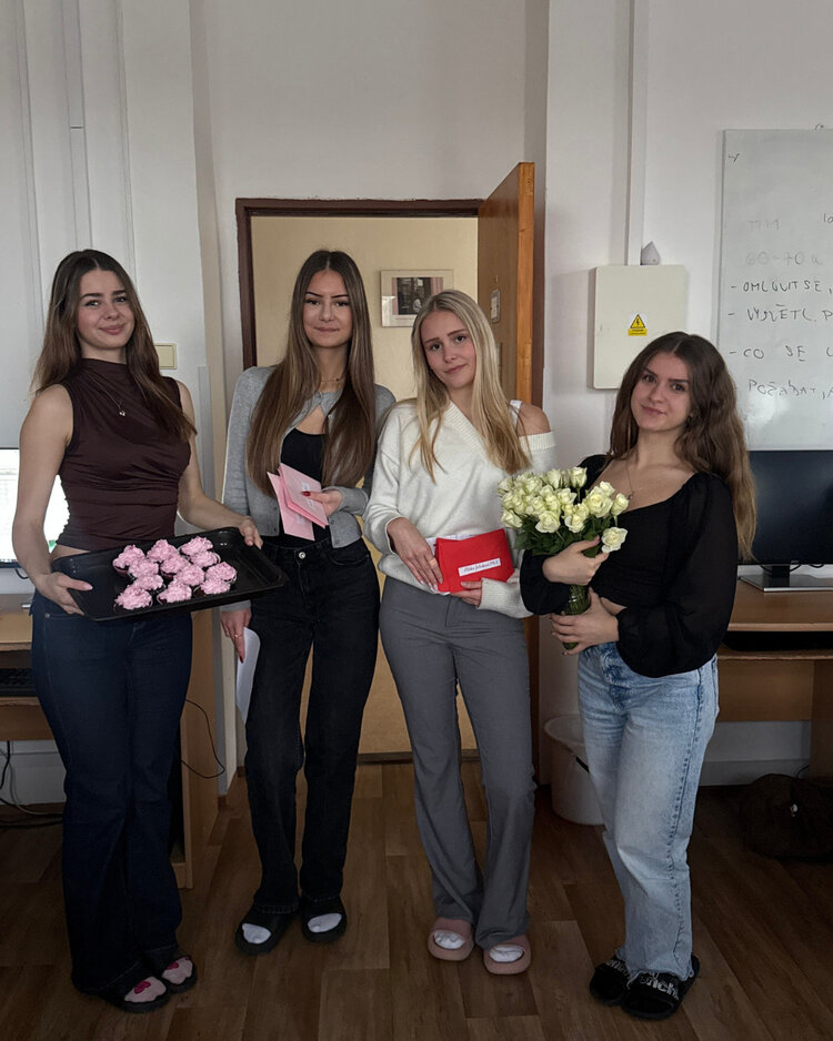 Valentinstag voller Freude und Hilfsbereitschaft Vier junge Frauen stehen nebeneinander in einem Raum. Die erste Frau hält ein Tablett mit rosa verzierten Cupcakes, die zweite und dritte halten jeweils eine Karte und ein kleines Geschenk, während die vierte Frau einen Strauß weißer Rosen trägt. Alle lächeln in die Kamera.