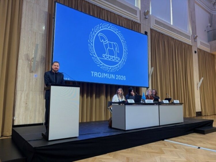 Modellsitzung der Vereinten Nationen TROJMUN 2026 Ein Mann in einem dunkelblauen Anzug spricht an einem Rednerpult auf einer Bühne. Im Hintergrund ist eine Leinwand mit dem Logo und dem Schriftzug „TROJMUN 2026“ zu sehen. Vier Personen sitzen an einem Tisch rechts auf der Bühne.
