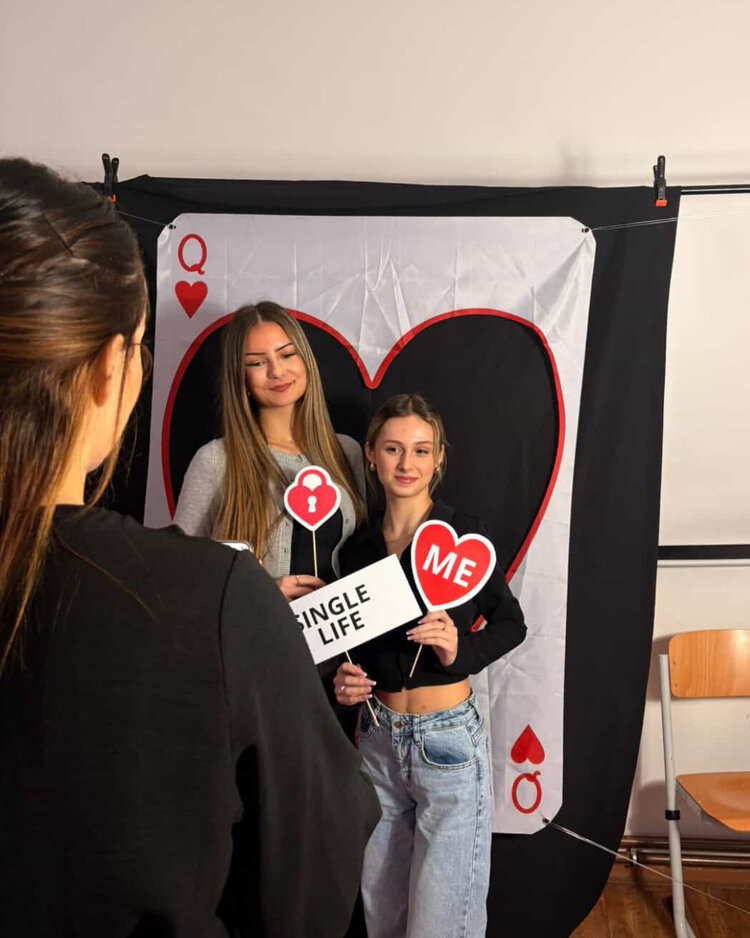 Valentinstag voller Freude und Hilfsbereitschaft Zwei junge Frauen posieren vor einem großen Spielkartenmotiv (Herz Queen). Eine hält ein Schild mit der Aufschrift "Single Life", die andere ein Schild mit einem Herz und der Aufschrift "ME". Eine Person im Vordergrund fotografiert sie.